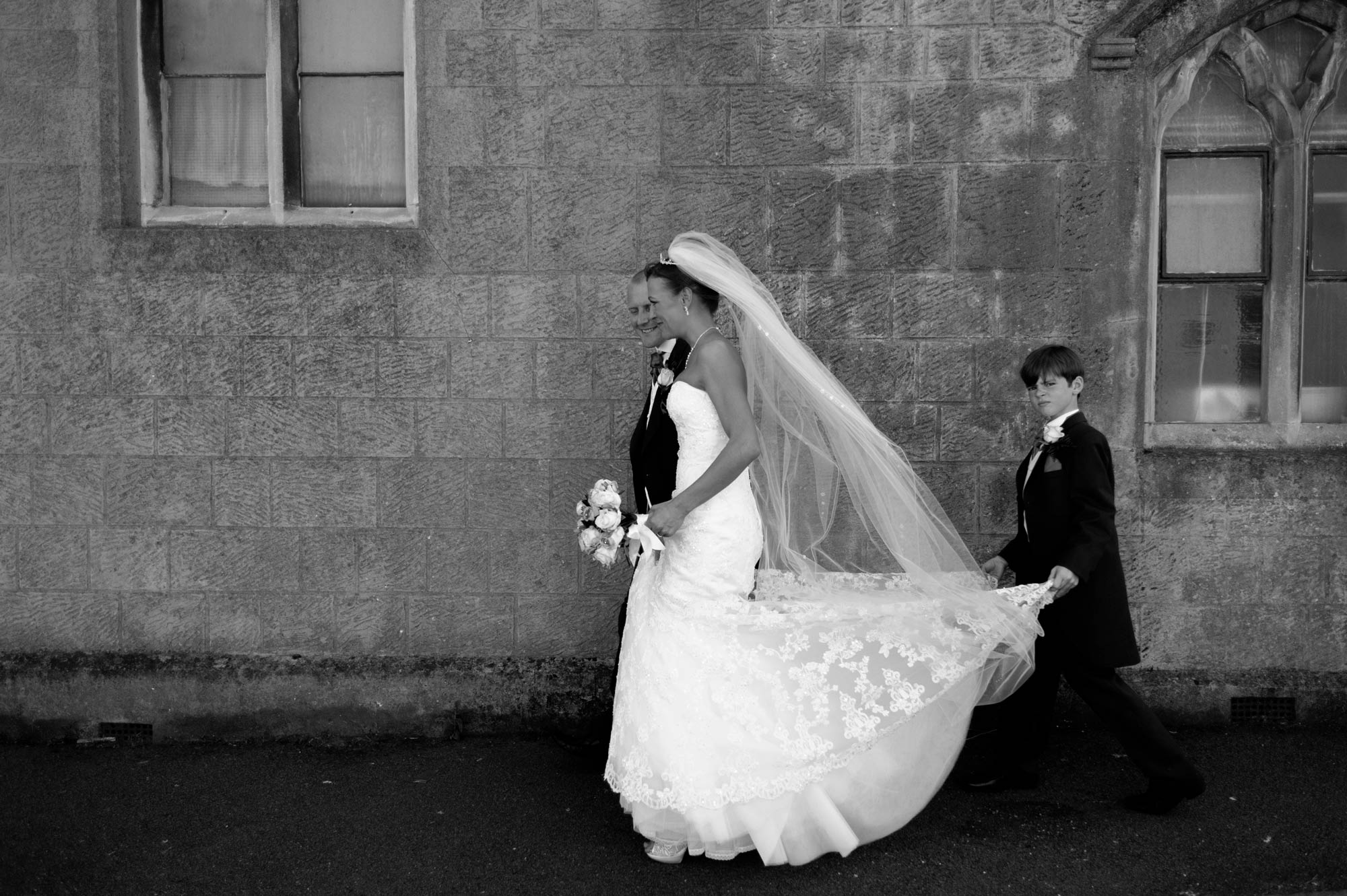 Pageboy holding bride's dress as she walks with groom